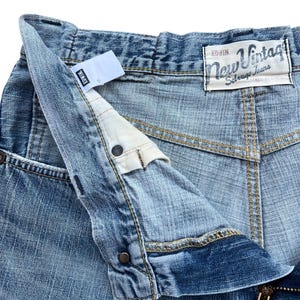 Pu&ograve; includere: Primo piano di jeans in denim blu chiaro con l'etichetta del marchio "EDWIN" visibile. I jeans presentano una chiusura con bottone e cerniera, con l'etichetta "New Vintage" sulla tasca posteriore. I jeans sono realizzati in denim resistente.
