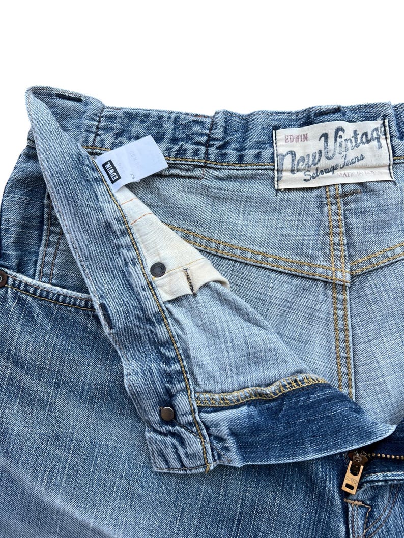Pu&ograve; includere: Primo piano di jeans in denim blu chiaro con l'etichetta del marchio "EDWIN" visibile. I jeans presentano una chiusura con bottone e cerniera, con l'etichetta "New Vintage" sulla tasca posteriore. I jeans sono realizzati in denim resistente.