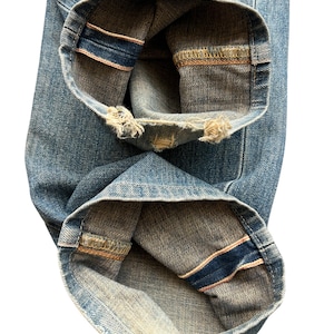 May include: Close-up of light blue denim jeans. The cuffs are rolled up, revealing a dark blue and pink selvedge edge. The denim has a worn appearance with frayed edges at the bottom of the cuffs.