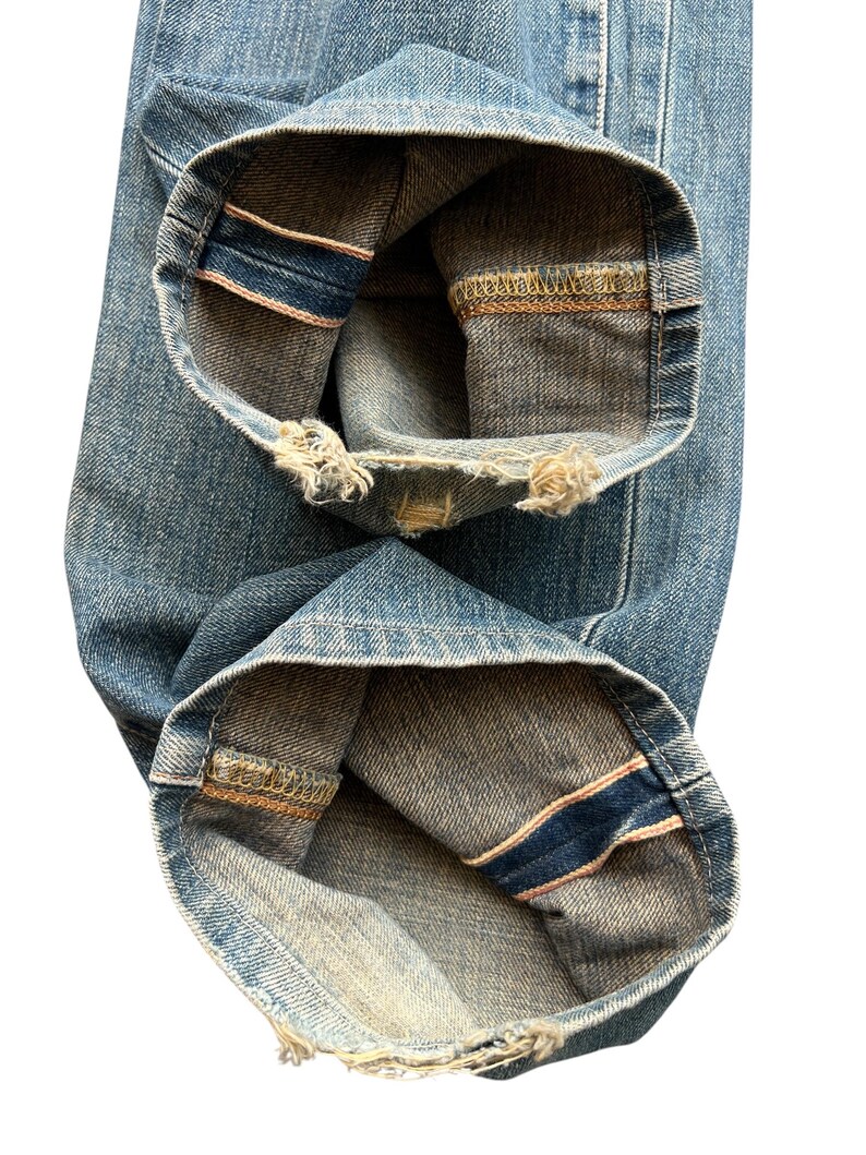 May include: Close-up of light blue denim jeans. The cuffs are rolled up, revealing a dark blue and pink selvedge edge. The denim has a worn appearance with frayed edges at the bottom of the cuffs.
