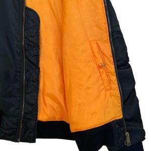 May include: Black bomber jacket with an orange lining. The jacket has a ribbed collar and cuffs, a zipper closure, and a pocket on the inside. The jacket is made of a durable material and is suitable for various weather conditions.