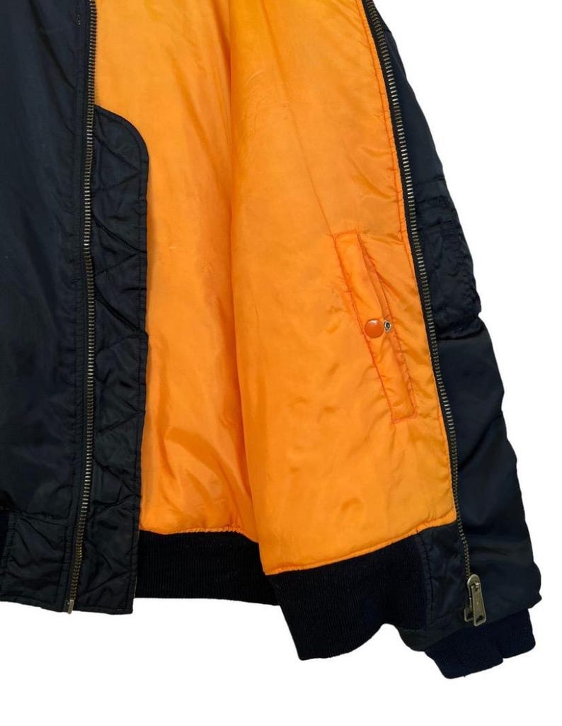 May include: Black bomber jacket with an orange lining. The jacket has a ribbed collar and cuffs, a zipper closure, and a pocket on the inside. The jacket is made of a durable material and is suitable for various weather conditions.