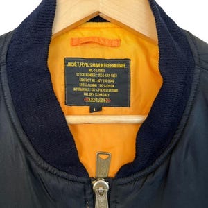 May include: Black bomber jacket with a navy blue collar and a bright orange lining. The jacket has a zipper closure and a label with text including "JACKET, FLVR'S MAN INTRERMEDIATE". The jacket is hanging on a wooden hanger.