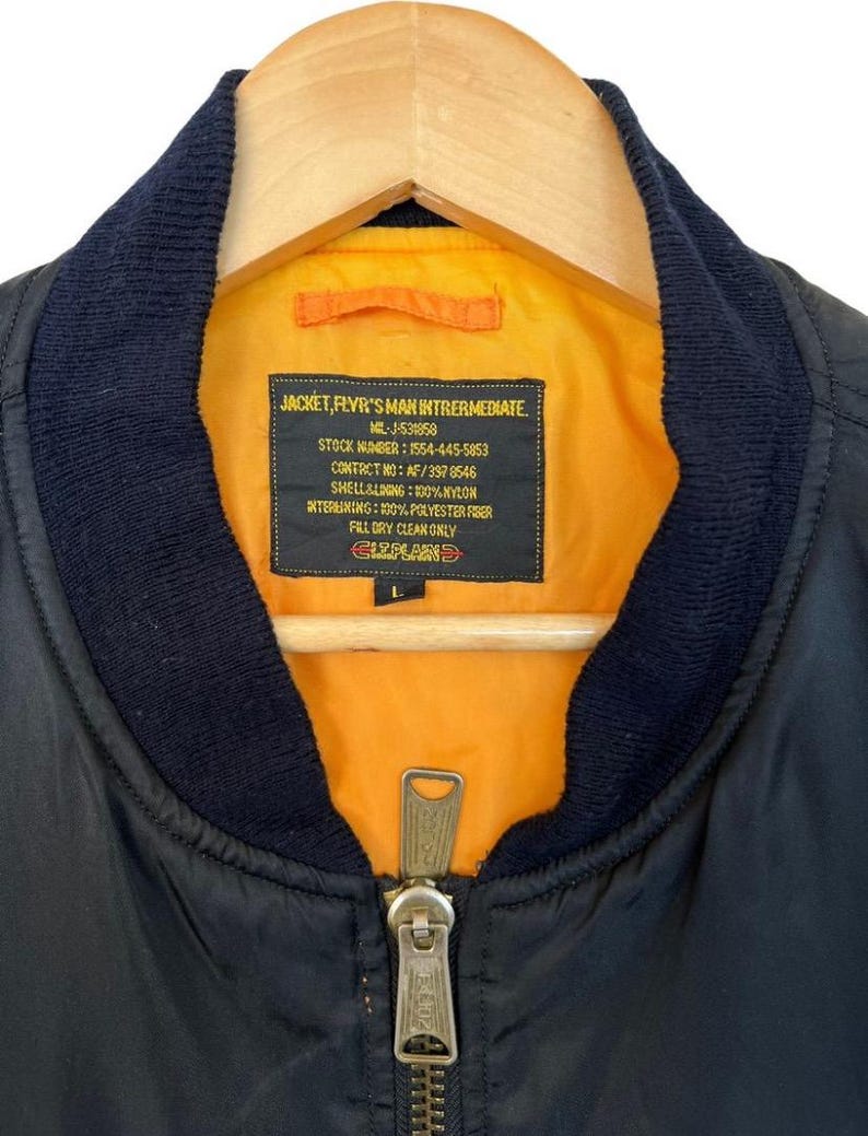 May include: Black bomber jacket with a navy blue collar and a bright orange lining. The jacket has a zipper closure and a label with text including "JACKET, FLVR'S MAN INTRERMEDIATE". The jacket is hanging on a wooden hanger.