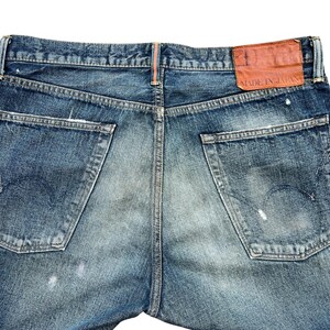 Puede incluir: Primer plano de la parte trasera de unos vaqueros azules con aspecto desgastado. Los vaqueros tienen dos bolsillos traseros, un parche de cuero marr&oacute;n con "MADE IN JAPAN" y salpicaduras de pintura blanca.