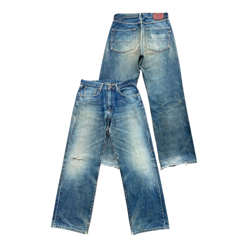 May include: Two pairs of faded blue denim jeans. One pair is laid flat, showing the front, with a distressed knee. The other pair is behind it, showing the back pockets and a red leather patch.