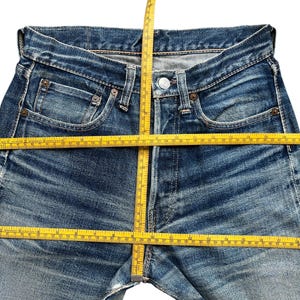 Pu&ograve; includere: Primo piano di jeans blu con un metro a nastro giallo posizionato sulla vita e sulle gambe. I jeans hanno una chiusura con bottoni, tasche e passanti per la cintura. Il denim ha un aspetto sbiadito e usurato.