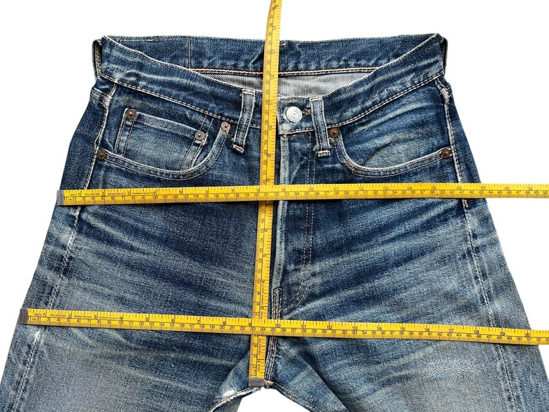 Pu&ograve; includere: Primo piano di jeans blu con un metro a nastro giallo posizionato sulla vita e sulle gambe. I jeans hanno una chiusura con bottoni, tasche e passanti per la cintura. Il denim ha un aspetto sbiadito e usurato.