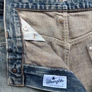 K&ouml;nnte beinhalten: Nahaufnahme einer blauen Jeans. Die Jeans hat einen Knopfverschluss und einen Rei&szlig;verschluss. Ein wei&szlig;es Etikett mit dem Wort "Wrangler" und "AUTHENTIC WESTERN WEAR" ist sichtbar.