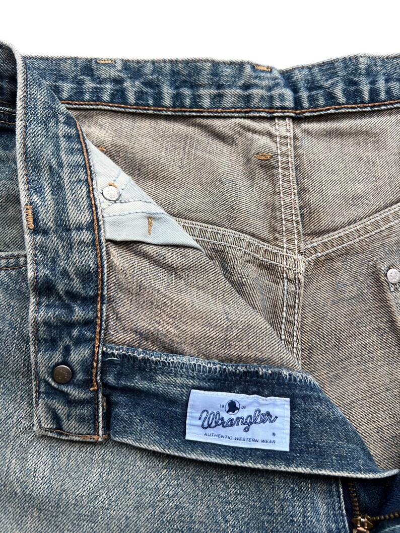 K&ouml;nnte beinhalten: Nahaufnahme einer blauen Jeans. Die Jeans hat einen Knopfverschluss und einen Rei&szlig;verschluss. Ein wei&szlig;es Etikett mit dem Wort "Wrangler" und "AUTHENTIC WESTERN WEAR" ist sichtbar.