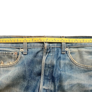 May include: Close-up of blue denim jeans with a yellow measuring tape across the waistband. The jeans have a classic five-pocket design and show signs of wear. The tape measures approximately 40 inches.