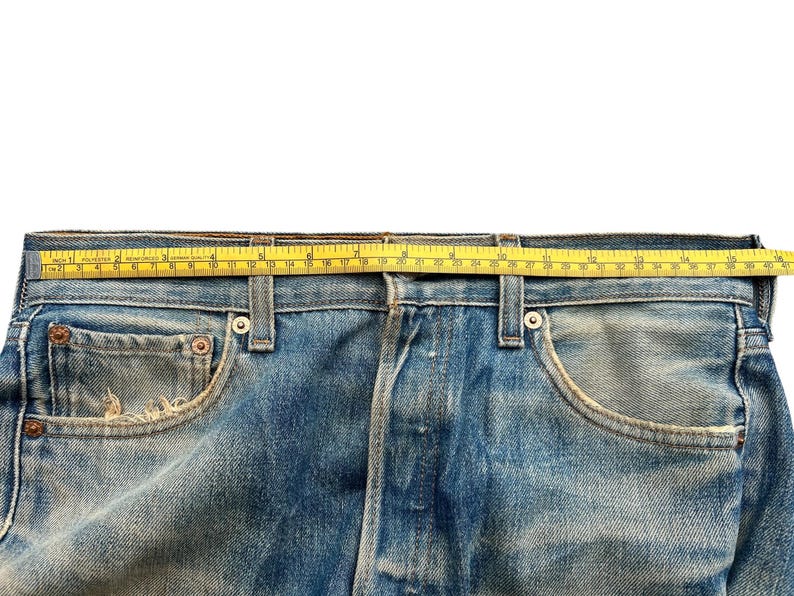 May include: Close-up of blue denim jeans with a yellow measuring tape across the waistband. The jeans have a classic five-pocket design and show signs of wear. The tape measures approximately 40 inches.
