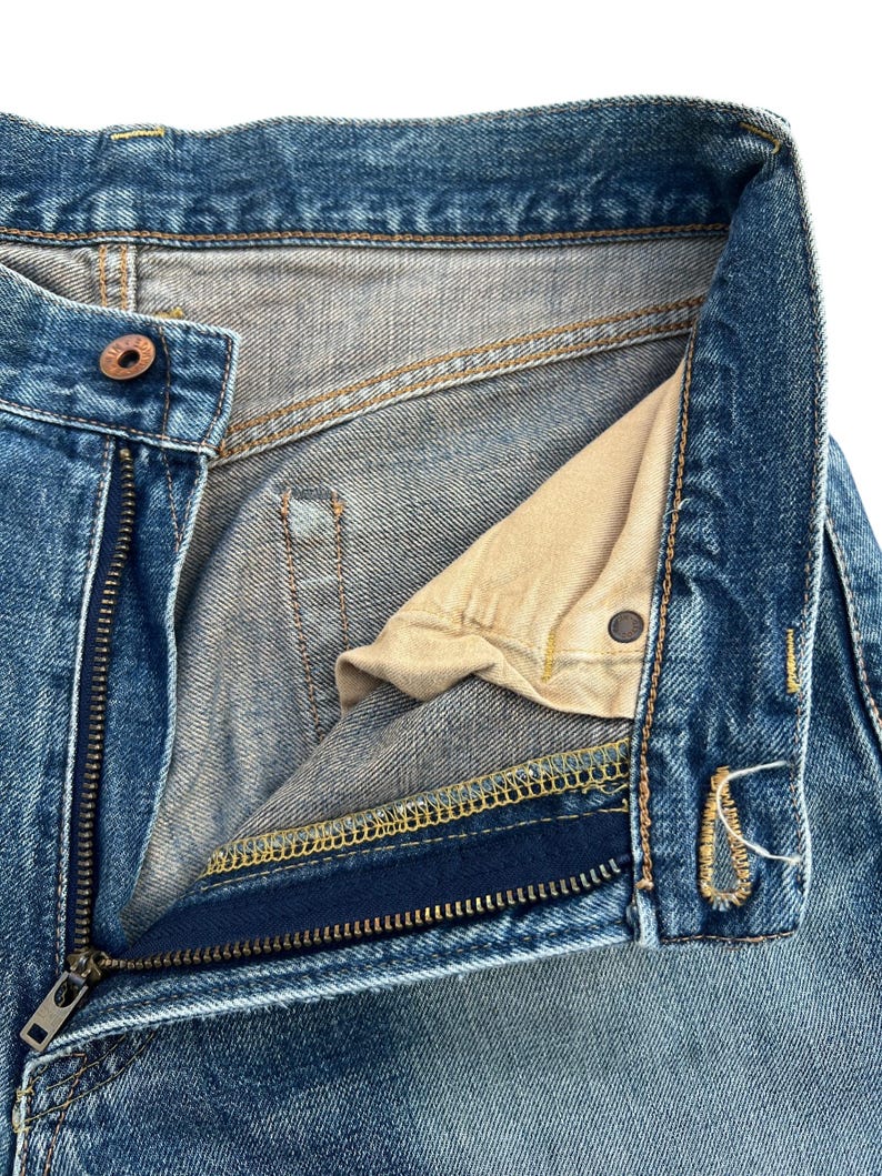 Puede incluir: Primer plano de la parte superior de unos vaqueros azules. La cremallera est&aacute; parcialmente desabrochada, revelando el forro interior y el cierre de bot&oacute;n. El denim tiene un aspecto descolorido y desgastado con costuras visibles.