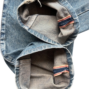 K&ouml;nnte beinhalten: Nahaufnahme der Manschetten einer hellblauen Jeans. Die Manschetten sind umgeschlagen und zeigen ein kontrastierendes graues Innenfutter und ein rot-wei&szlig;-blaues Streifendetail. Der Denim hat ein abgenutztes Aussehen.