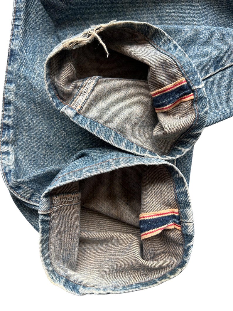 K&ouml;nnte beinhalten: Nahaufnahme der Manschetten einer hellblauen Jeans. Die Manschetten sind umgeschlagen und zeigen ein kontrastierendes graues Innenfutter und ein rot-wei&szlig;-blaues Streifendetail. Der Denim hat ein abgenutztes Aussehen.