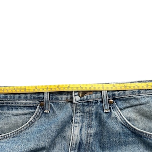 K&ouml;nnte beinhalten: Nahaufnahme einer hellblauen Jeans mit einem gelben Ma&szlig;band oben. Die Jeans hat einen klassischen F&uuml;nf-Taschen-Stil mit Metallnieten und einem Knopfverschluss. Das Ma&szlig;band zeigt eine L&auml;nge von etwa 48 cm.