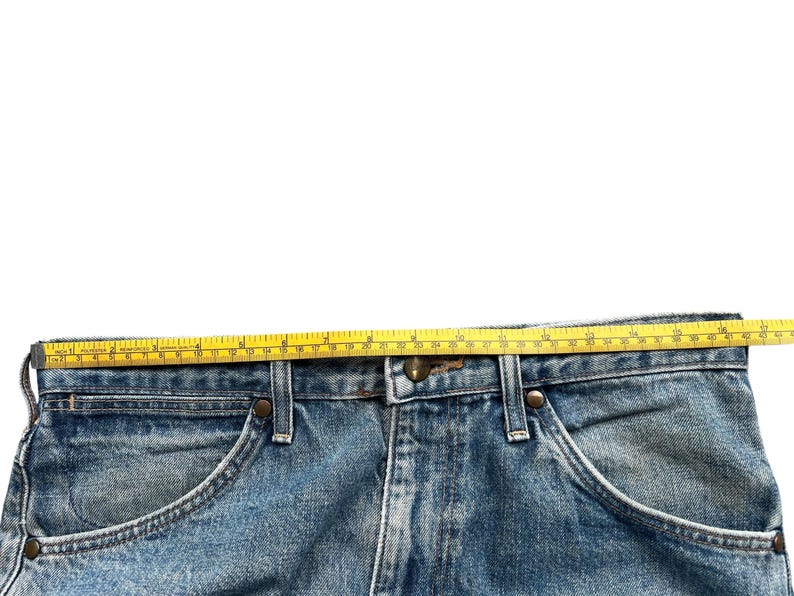 K&ouml;nnte beinhalten: Nahaufnahme einer hellblauen Jeans mit einem gelben Ma&szlig;band oben. Die Jeans hat einen klassischen F&uuml;nf-Taschen-Stil mit Metallnieten und einem Knopfverschluss. Das Ma&szlig;band zeigt eine L&auml;nge von etwa 48 cm.