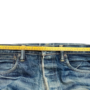 May include: Close-up of blue denim jeans with a yellow measuring tape across the waist. The jeans have classic five-pocket styling and visible stitching. The tape measures the waist width in inches.