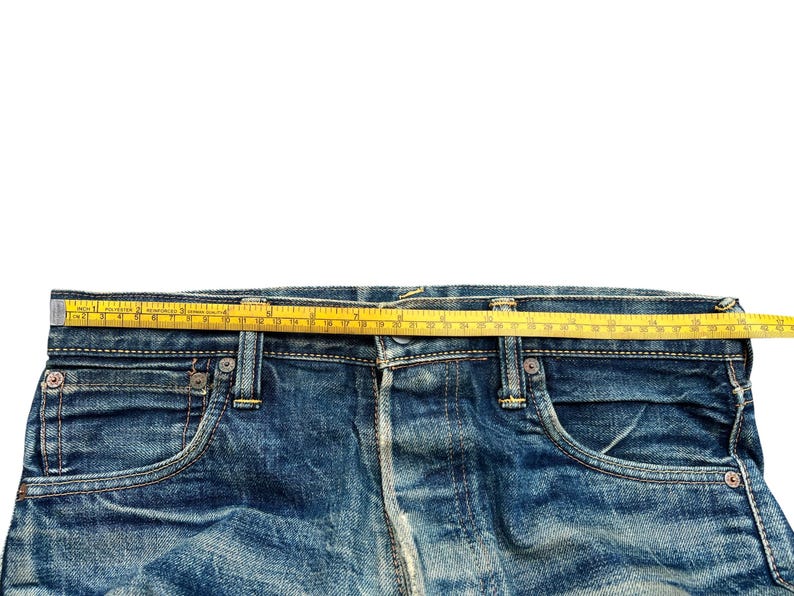 May include: Close-up of blue denim jeans with a yellow measuring tape across the waist. The jeans have classic five-pocket styling and visible stitching. The tape measures the waist width in inches.