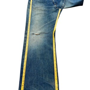 May include: Faded blue denim jeans with a distressed knee area. A yellow measuring tape is placed along the length of the jeans. The jeans are folded over at the top.