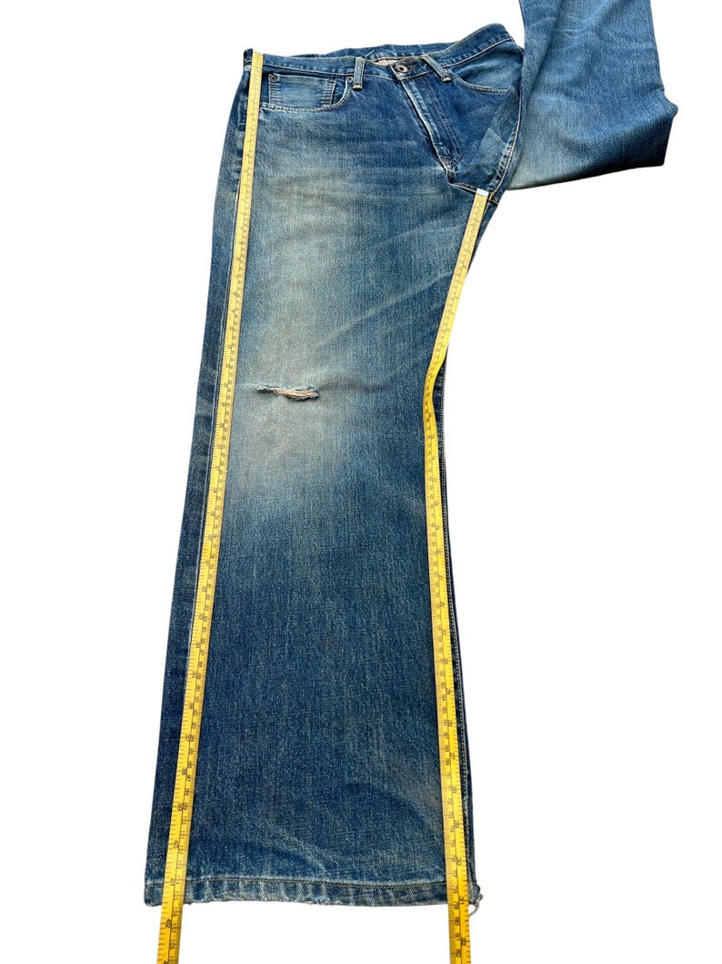 May include: Faded blue denim jeans with a distressed knee area. A yellow measuring tape is placed along the length of the jeans. The jeans are folded over at the top.