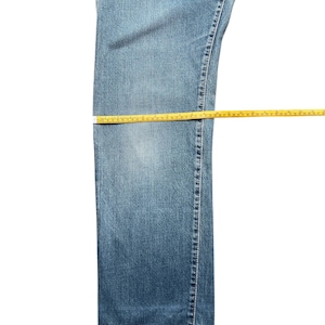 May include: Blue denim jeans with yellow measuring tape laid across the waist, thigh, and hem. The jeans are a medium wash and appear to be new. The measuring tape is in inches.