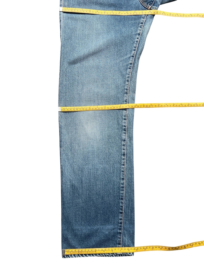 May include: Blue denim jeans with yellow measuring tape laid across the waist, thigh, and hem. The jeans are a medium wash and appear to be new. The measuring tape is in inches.