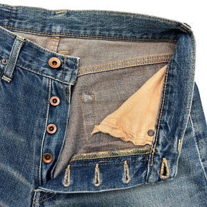 Puede incluir: Primer plano de unos vaqueros de mezclilla azules con bragueta de botones. Los vaqueros muestran signos de desgaste y decoloraci&oacute;n, con costuras visibles y botones color cobre. El forro interior del bolsillo es de color beige claro.
