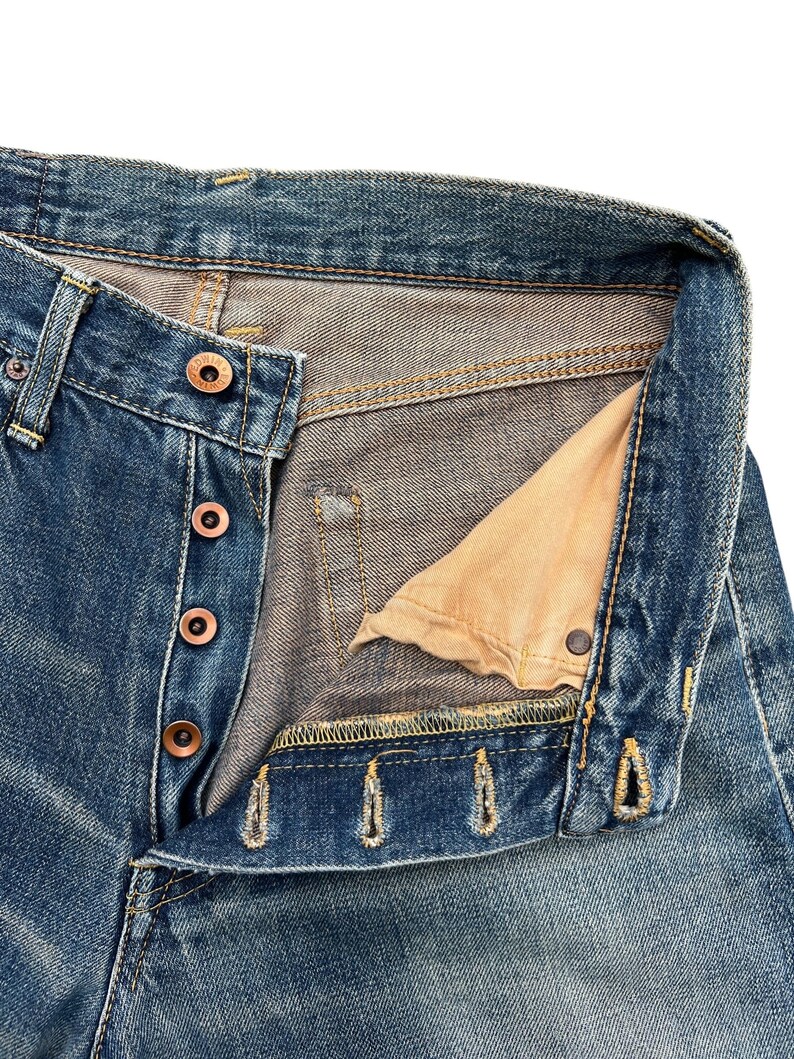 Puede incluir: Primer plano de unos vaqueros de mezclilla azules con bragueta de botones. Los vaqueros muestran signos de desgaste y decoloraci&oacute;n, con costuras visibles y botones color cobre. El forro interior del bolsillo es de color beige claro.