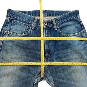 May include: Blue denim jeans with a classic five-pocket design. A yellow measuring tape is laid across the jeans, indicating measurements. The jeans have a button and zipper closure and show signs of wear.