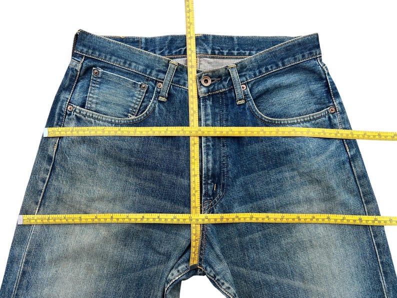 May include: Blue denim jeans with a classic five-pocket design. A yellow measuring tape is laid across the jeans, indicating measurements. The jeans have a button and zipper closure and show signs of wear.