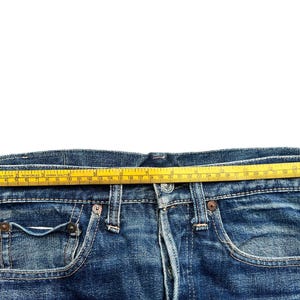 Pu&ograve; includere: Primo piano di jeans blu con un metro a nastro giallo steso sulla vita. Il nastro mostra le misure in centimetri. I jeans hanno un classico stile a cinque tasche e una chiusura con bottoni.