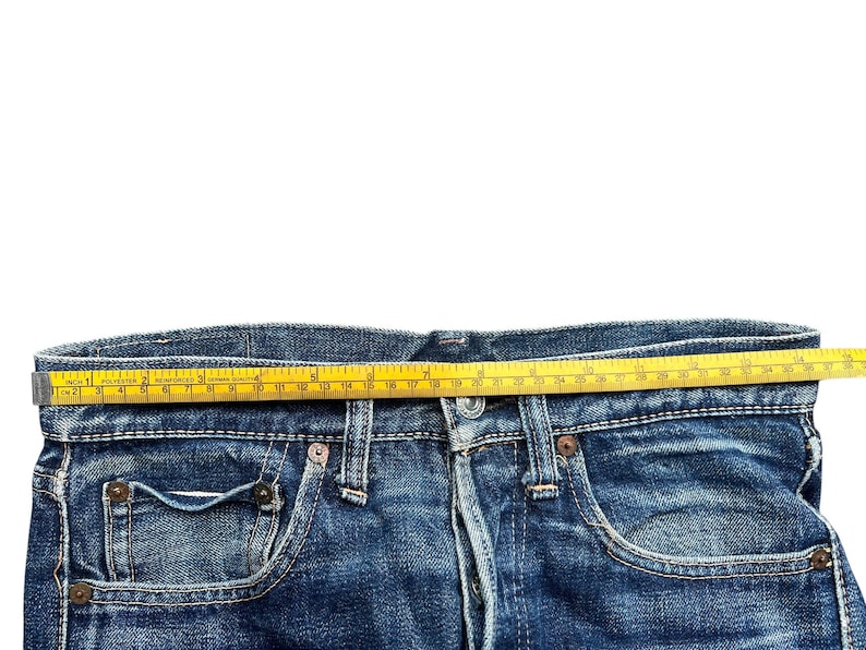 Pu&ograve; includere: Primo piano di jeans blu con un metro a nastro giallo steso sulla vita. Il nastro mostra le misure in centimetri. I jeans hanno un classico stile a cinque tasche e una chiusura con bottoni.