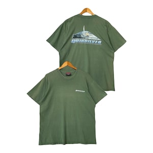 May include: Two olive green t-shirts on wooden hangers. One shirt displays the Quiksilver logo on the front and a graphic on the back. The other shirt has the Quiksilver logo on the front. Both shirts have short sleeves and a crew neck.