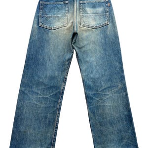 May include: Back view of faded blue denim jeans with two back pockets. The jeans have a worn appearance with visible wear marks and a leather patch at the waistband. The jeans have a straight leg cut.