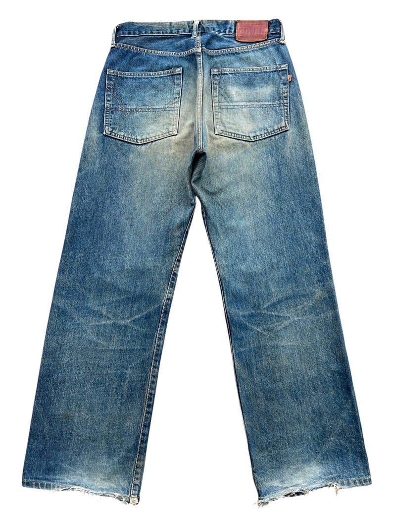 May include: Back view of faded blue denim jeans with two back pockets. The jeans have a worn appearance with visible wear marks and a leather patch at the waistband. The jeans have a straight leg cut.