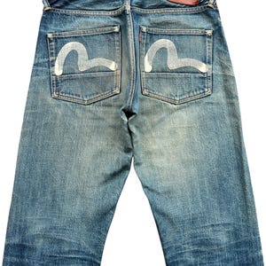 May include: Faded blue denim jeans with white painted design on the back pockets. The jeans have a red leather patch and a classic five-pocket design. The denim shows signs of wear and fading.