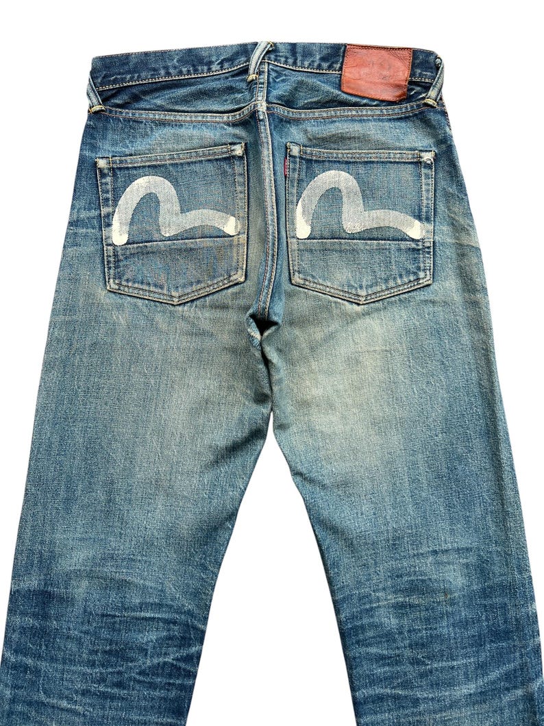 May include: Faded blue denim jeans with white painted design on the back pockets. The jeans have a red leather patch and a classic five-pocket design. The denim shows signs of wear and fading.