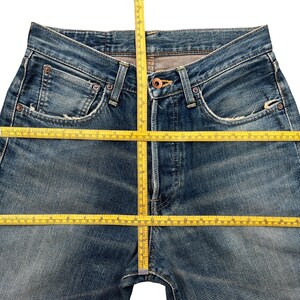 Puede incluir: Primer plano de unos vaqueros azules con una cinta m&eacute;trica amarilla colocada sobre la cintura y la entrepierna. Los vaqueros tienen un dise&ntilde;o cl&aacute;sico de cinco bolsillos, con desgaste y decoloraci&oacute;n visibles. El denim es de lavado medio.