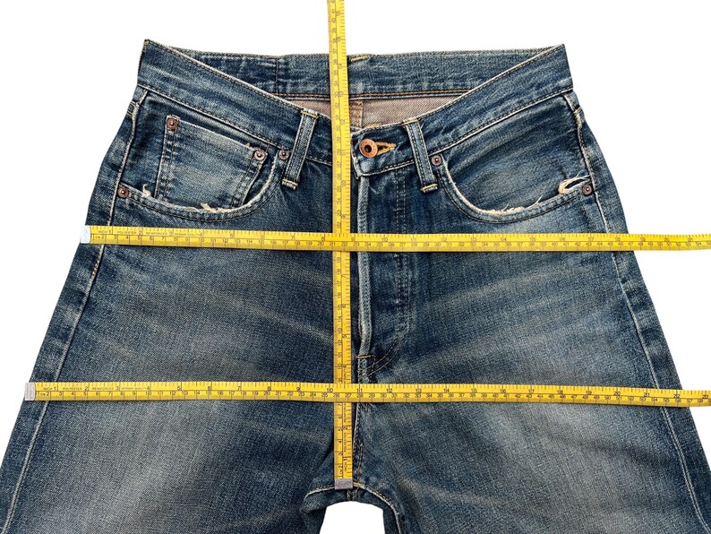 Puede incluir: Primer plano de unos vaqueros azules con una cinta m&eacute;trica amarilla colocada sobre la cintura y la entrepierna. Los vaqueros tienen un dise&ntilde;o cl&aacute;sico de cinco bolsillos, con desgaste y decoloraci&oacute;n visibles. El denim es de lavado medio.