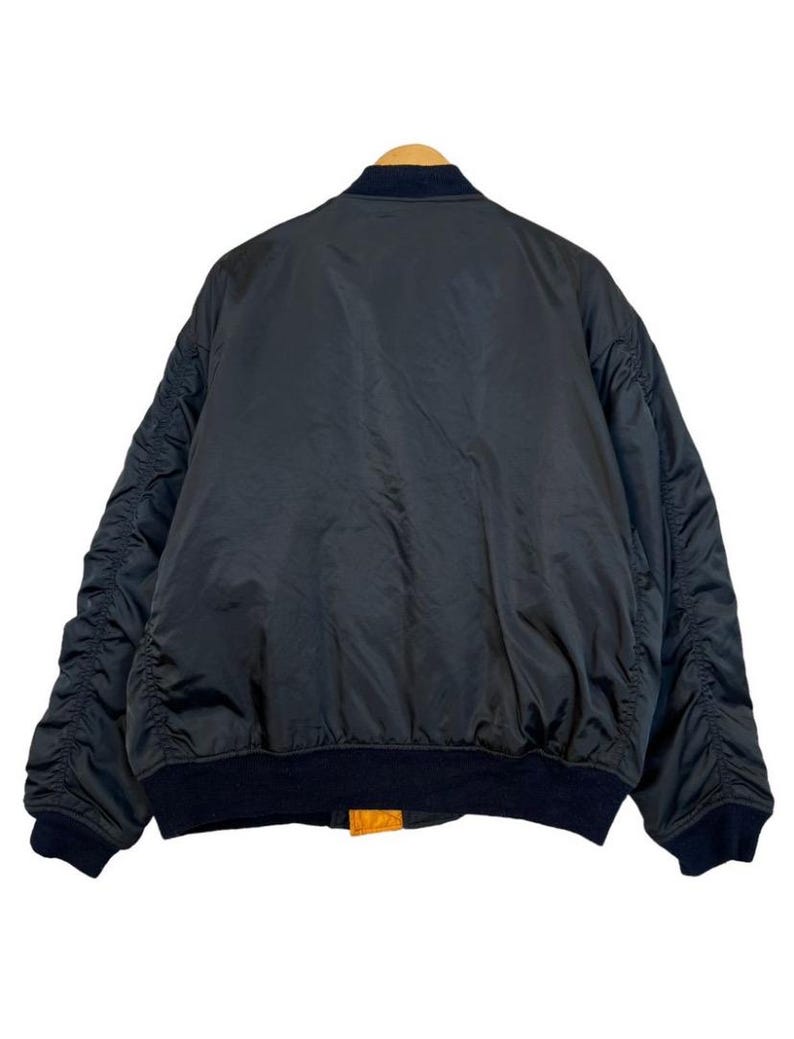 May include: A black bomber jacket with a navy blue collar and cuffs. The jacket has a smooth, shiny texture and a ribbed hem. The jacket is hanging on a wooden hanger.