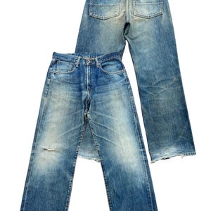 May include: Two pairs of blue denim jeans. One pair is laid on top of the other. The jeans have a faded wash, distressed details, and classic five-pocket styling. The front pair has a small tear near the knee.