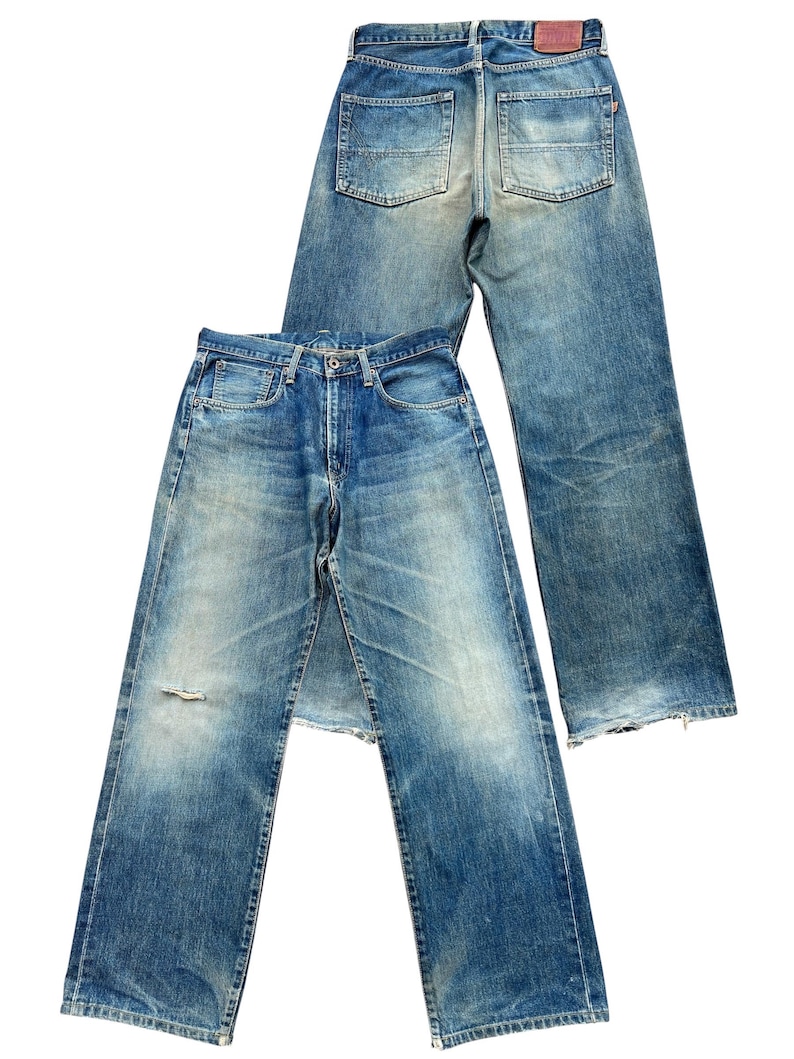 May include: Two pairs of blue denim jeans. One pair is laid on top of the other. The jeans have a faded wash, distressed details, and classic five-pocket styling. The front pair has a small tear near the knee.
