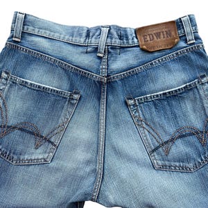Pu&ograve; includere: Primo piano del retro di jeans di denim blu chiaro. I jeans hanno due tasche posteriori con cuciture decorative e un'etichetta in pelle marrone con il marchio "EDWIN". I jeans sono sbiaditi e hanno un aspetto usurato.