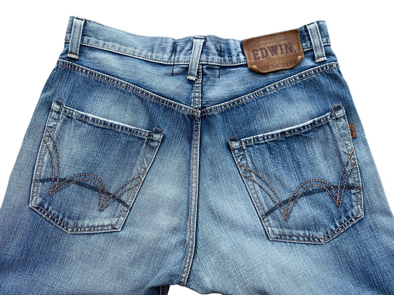 Pu&ograve; includere: Primo piano del retro di jeans di denim blu chiaro. I jeans hanno due tasche posteriori con cuciture decorative e un'etichetta in pelle marrone con il marchio "EDWIN". I jeans sono sbiaditi e hanno un aspetto usurato.
