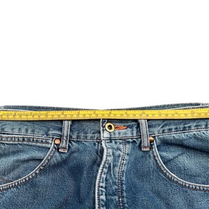 May include: Close-up of blue denim jeans with a yellow measuring tape laid across the waistband. The jeans have classic five-pocket styling with orange stitching and metal rivets. The tape measures the waist of the jeans.