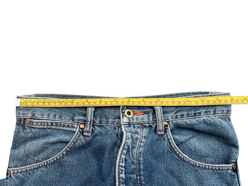 May include: Close-up of blue denim jeans with a yellow measuring tape laid across the waistband. The jeans have classic five-pocket styling with orange stitching and metal rivets. The tape measures the waist of the jeans.