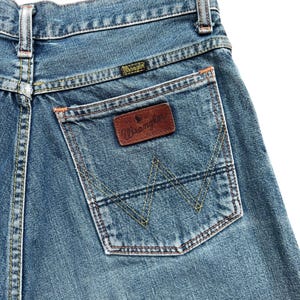 May include: Close-up of the back of blue denim jeans. The jeans feature a back pocket with a brown leather patch and the Wrangler logo. The pocket has a distinctive stitched design. The jeans have orange and yellow stitching.