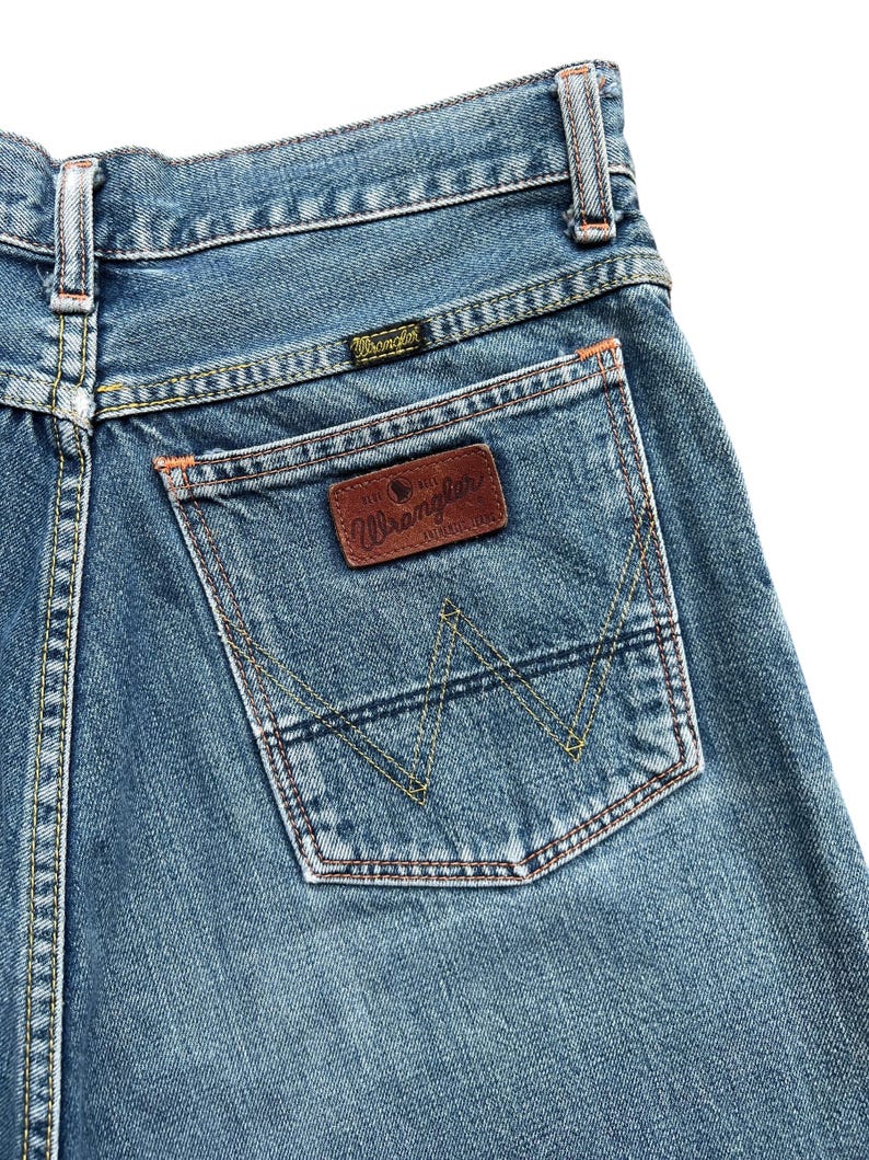 May include: Close-up of the back of blue denim jeans. The jeans feature a back pocket with a brown leather patch and the Wrangler logo. The pocket has a distinctive stitched design. The jeans have orange and yellow stitching.