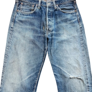 以下が含まれることがあります： ボタンフライとクラシックな5ポケットデザインの、色あせたブルーデニムジーンズ。右脚に小さな破れなど、使用感があり、ヴィンテージ感のあるジーンズです。